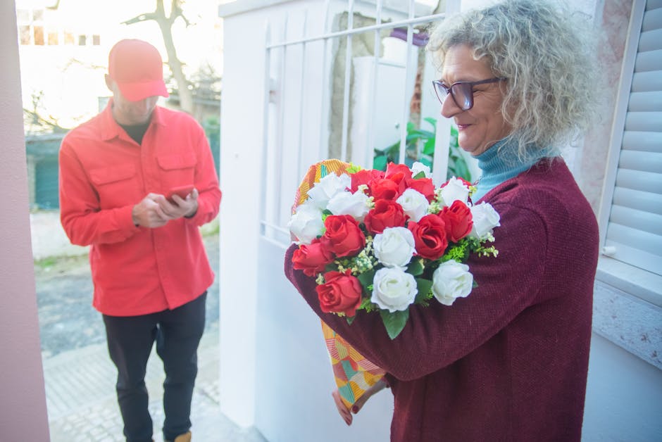 A woman with curly gray hair, wearing glasses, a blue turtleneck, and a maroon sweater, is holding a vibrant bouquet of fresh red and white roses, accented with green foliage, wrapped in colorful pape