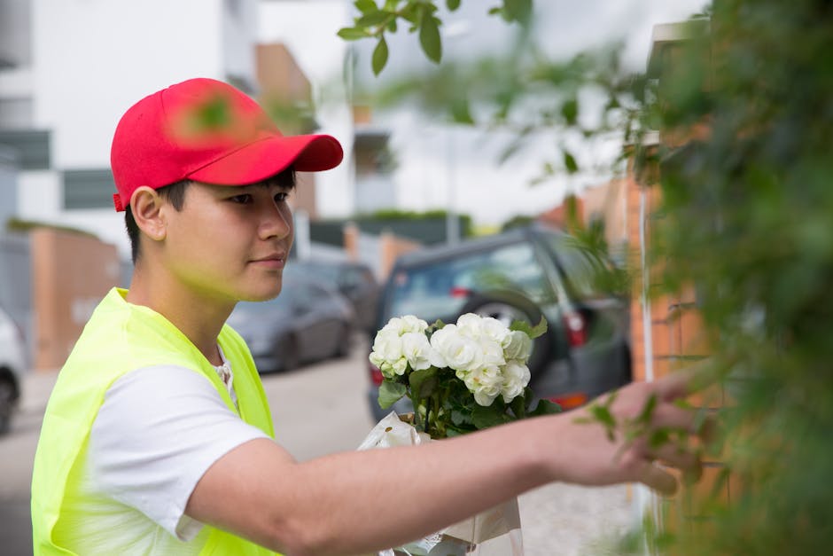 A young delivery person wearing a red cap and high-visibility yellow vest is shown in an outdoor urban setting, delivering a floral arrangement. The bouquet consists of fresh white hydrangeas with lus