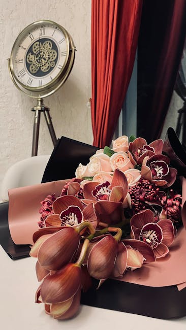 A floral arrangement of deep burgundy orchids and soft pink roses, stylishly wrapped in pink and brown paper with a satin ribbon, displayed on a dark surface in an indoor setting. The arrangement show