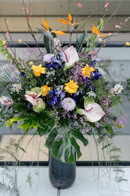 A large, vibrant floral arrangement in a tall, dark vase featuring a variety of fresh flowers including yellow daisies, purple asters, birch branches with pink blossoms, white calla lilies with pink s