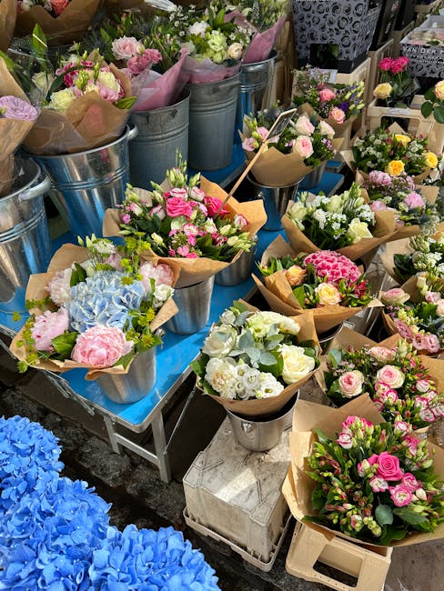 A vibrant display of floral bouquets arranged in metal buckets and paper wraps on a table, featuring an assortment of fresh flowers including pink roses, white and pink peonies, light blue hydrangeas,