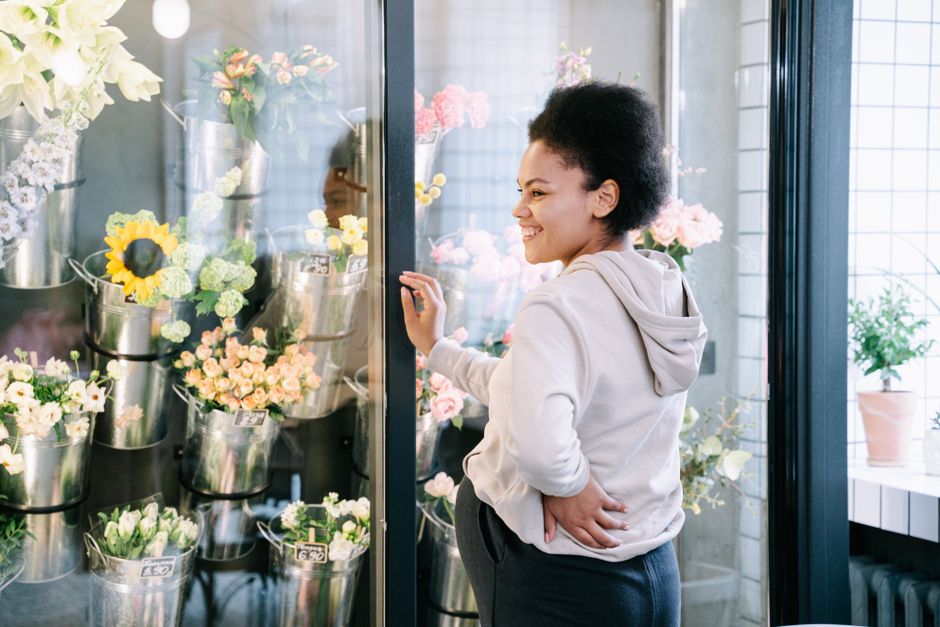 A young woman with curly black hair, smiling and dressed in a light grey hoodie, is standing inside a flower shop, [flowersfulham.com], near a glass display case filled with various fresh flower arran