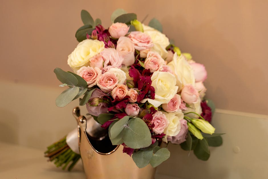 A floral arrangement featuring a bouquet of fresh flowers, including pale pink roses, white roses, and deep burgundy blooms, with prominent eucalyptus leaves interspersed throughout. The bouquet is he